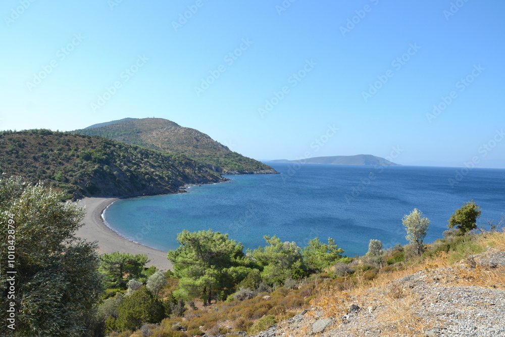 The photo reveals a stunning view of the Aegean Sea's coastline, with crystal blue waters meeting a rugged shoreline. In the background, majestic mountains rise against the clear sky, creating a breat