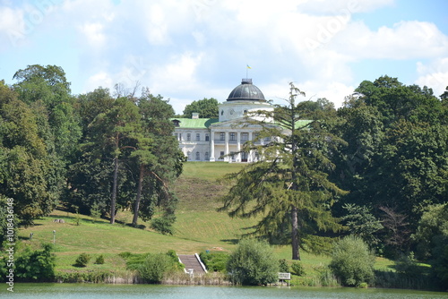 The photo shows the Kachanivka estate in summer, with its grand neoclassical mansion surrounded by lush greenery. A well-manicured lawn and vibrant trees frame the bright, sunlit building. 