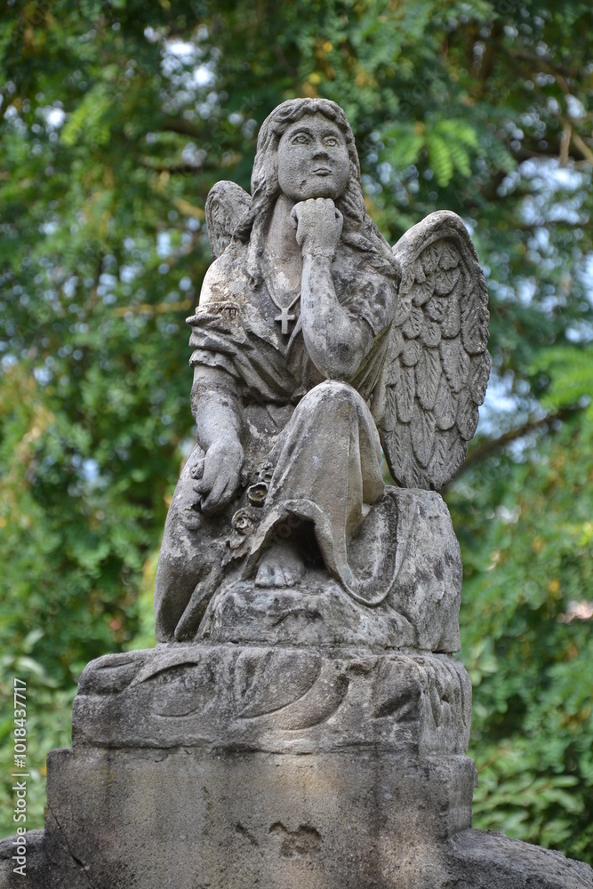 The image depicts a stone statue of an angel sitting thoughtfully, resting her chin on her hand. The angel has detailed wings, flowing garments, and a serene expression, surrounded by greenery. The image depicts a stone statue of an angel sitting thoughtfully, resting her chin on her hand. The angel has detailed wings, flowing garments, and a serene expression, surrounded by greenery.