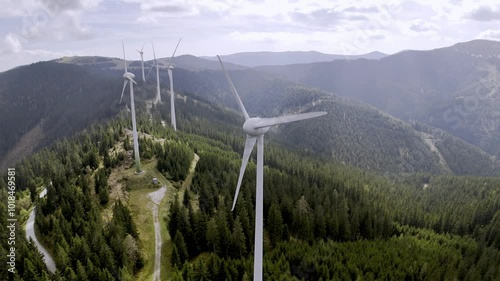 Wind turbines on a forested mountain, set against a bright sky with clouds, representing renewable energy in nature.
