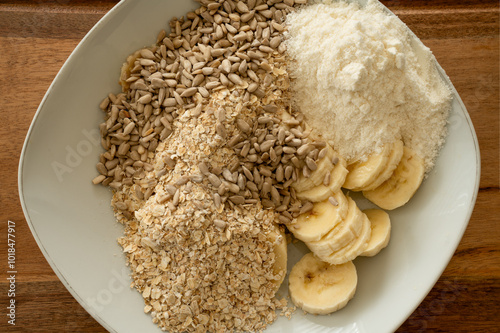 Close up of the preparation of a deep plate with muesli ingredients, starting with oatmeal, sunflower seeds, banana slices, protein powder. Table stands on a wooden snackboard. photo