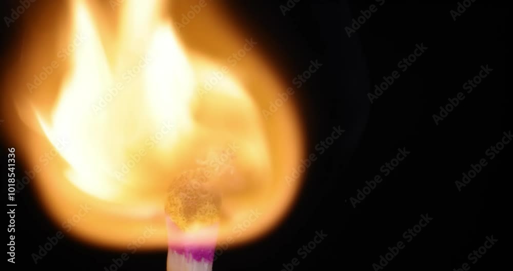 A close-up of a lit match, showcasing a vibrant yellow-orange flame against a dark background. The match head is colored pink. A close-up of a lit match, showcasing a vibrant yellow-orange flame against a dark background. The match head is colored pink.