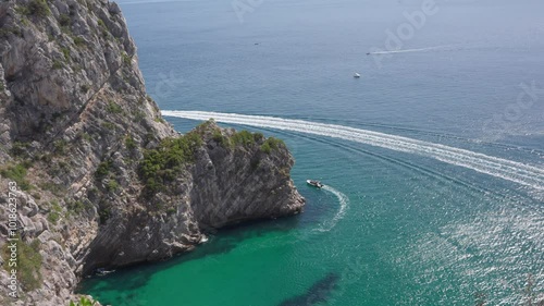 The scene is set against a steep rocky cliff adorned with sparse greenery, while the boats create a dynamic contrast against the calm sea.