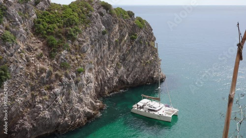 The image captures a serene coastal scene featuring a sailboat moored near a rugged rocky cliff. The turquoise water contrasts with the towering rock formation covered in patches of greenery.