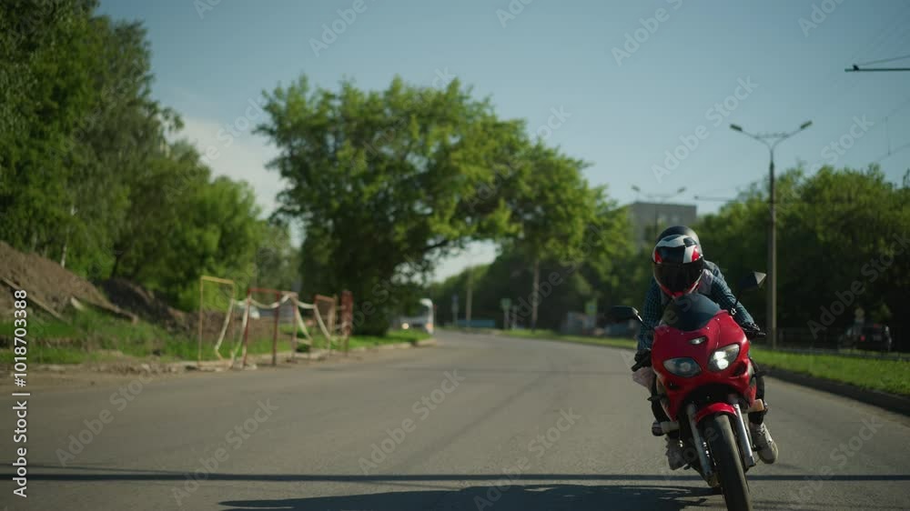 Two friends ride a red power bike, both wearing helmets, navigating the road with parked cars along the roadside, electric poles and lush trees are visible in the background Two friends ride a red power bike, both wearing helmets, navigating the road with parked cars along the roadside, electric poles and lush trees are visible in the background