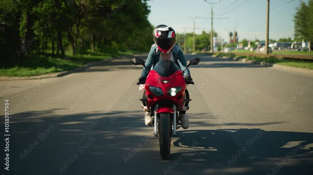 Two friends ride a power bike, both wearing helmets, along a shaded tarred road, tree shadows cast across the road, while cars are seen moving in the opposite lane Two friends ride a power bike, both wearing helmets, along a shaded tarred road, tree shadows cast across the road, while cars are seen moving in the opposite lane