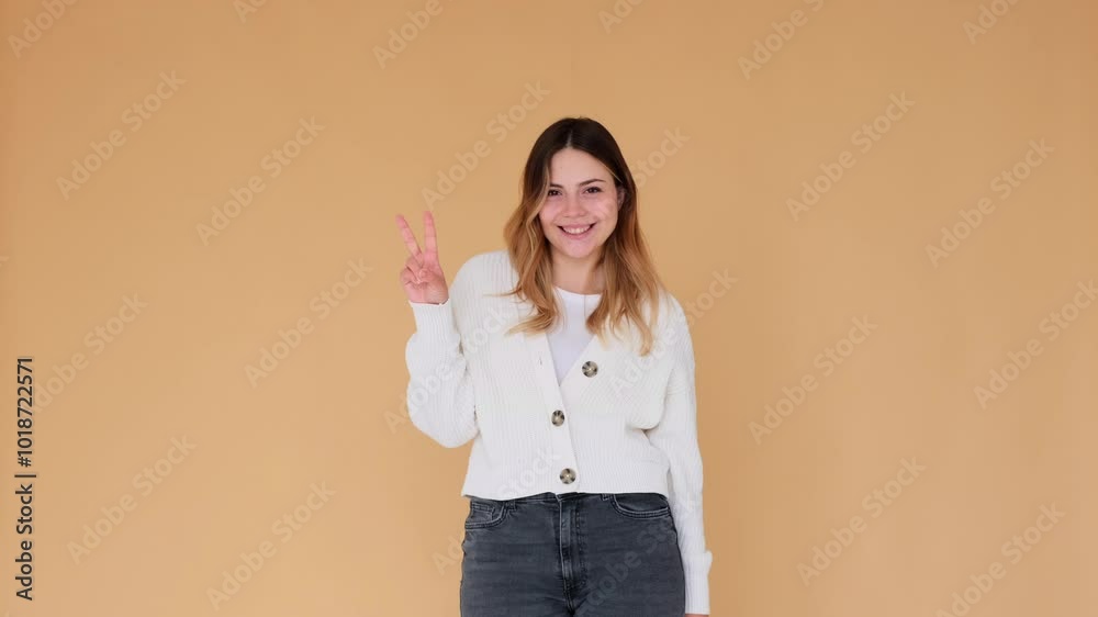 Excited and smiley Caucasian young adult woman showing victory or peace sign, posing on beige background. Accomplishment and triumph symbol. Excited and smiley Caucasian young adult woman showing victory or peace sign, posing on beige background. Accomplishment and triumph symbol.