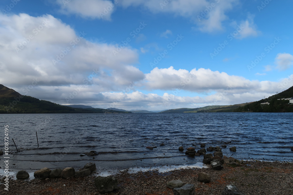 A serene Scottish loch scene with a rocky shoreline, surrounded by hills and forests under a partly cloudy sky. The water is slightly rippled, and the horizon shows distant hills and scattered clouds. A serene Scottish loch scene with a rocky shoreline, surrounded by hills and forests under a partly cloudy sky. The water is slightly rippled, and the horizon shows distant hills and scattered clouds.