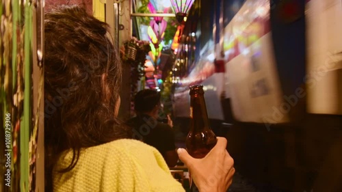 Excited caucasian woman stand by rails drink alcohol and watch passing train in famous Hanoi train street in Hanoi city, capital Vietnam. Nightlife concept
