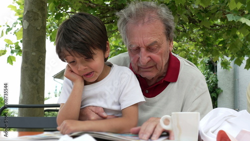 Elderly man and young boy reading a book together outdoors, with the boy showing a puzzled expression as they share a moment of learning and bonding under the shade of a tree