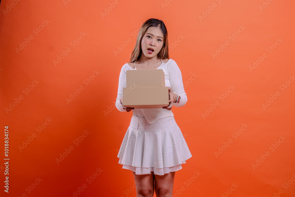 Fototapeta premium young asian woman in white top surprised to camera both hands holding 2 plain cardboard boxes, standing on orange background. for delivery concept, gift, expedition, service, package