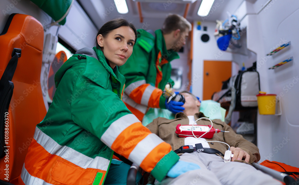Paramedics in green and orange uniforms tend to male patient inside an ambulance. One monitors the patient while another provides oxygen. The scene highlights emergency medical response and teamwork Paramedics in green and orange uniforms tend to male patient inside an ambulance. One monitors the patient while another provides oxygen. The scene highlights emergency medical response and teamwork