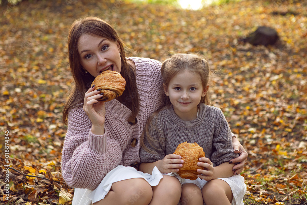 mother and her daughter of seven years walk warm autumn in a park with trees at sunset in orange leaves. sitting on the ground eating croissants.