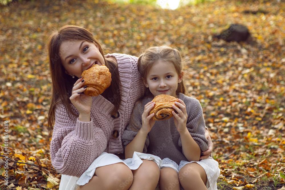 mother and her daughter of seven years walk warm autumn in a park with trees at sunset in orange leaves. sitting on the ground eating croissants.