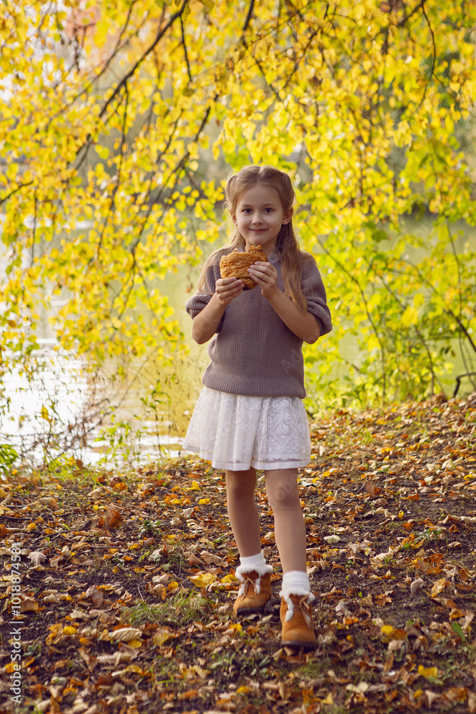 A girl of seven holds a curroissant bun in the park at sunset in autumn