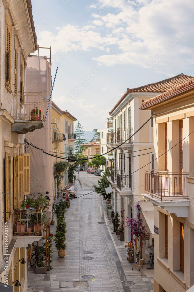 Naklejka premium Wander through a picturesque Nafplio street adorned with vibrant blooms and inviting balconies, leading toward a serene coastal view illuminated by the midday sun.