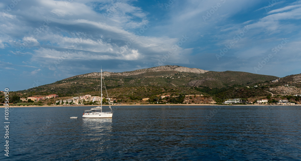 Fototapeta premium Sailboat sailing on a vast blue sea on a sunny day with cloudy sky near shore of Greek island
