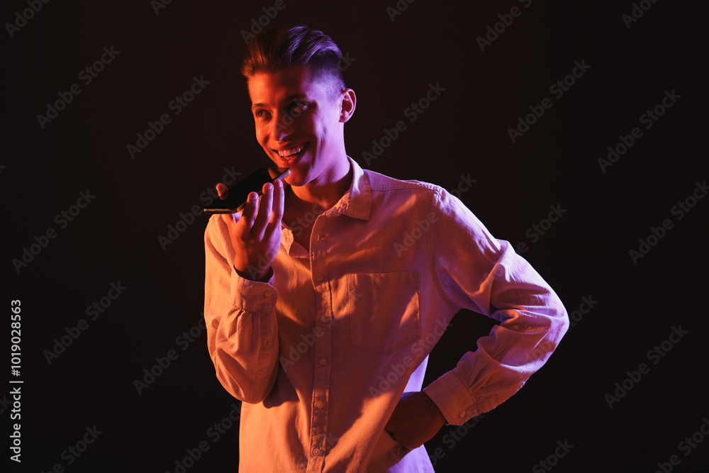 Young man recording voice message via smartphone on dark background with neon lights Young man recording voice message via smartphone on dark background with neon lights