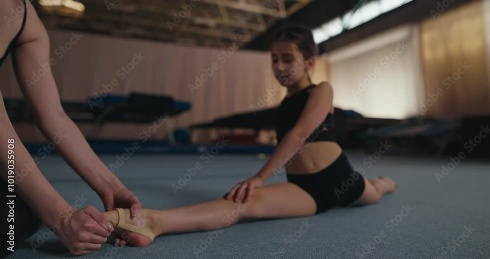 Close up a little girl gymnast in a sports uniform sits on the cross splits and learns how to pull her toe correctly with the help of a girl trainer in the gym