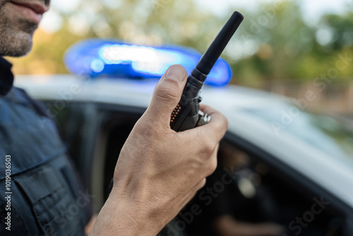 Police officer using walkie talkie near patrol car with flashing lights
