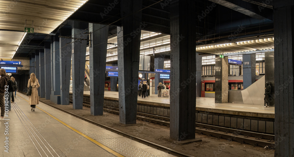 A modern underground train station with passengers waiting on platforms, illuminated by artificial light. People stand near the tracks, and a train is visible in the background.