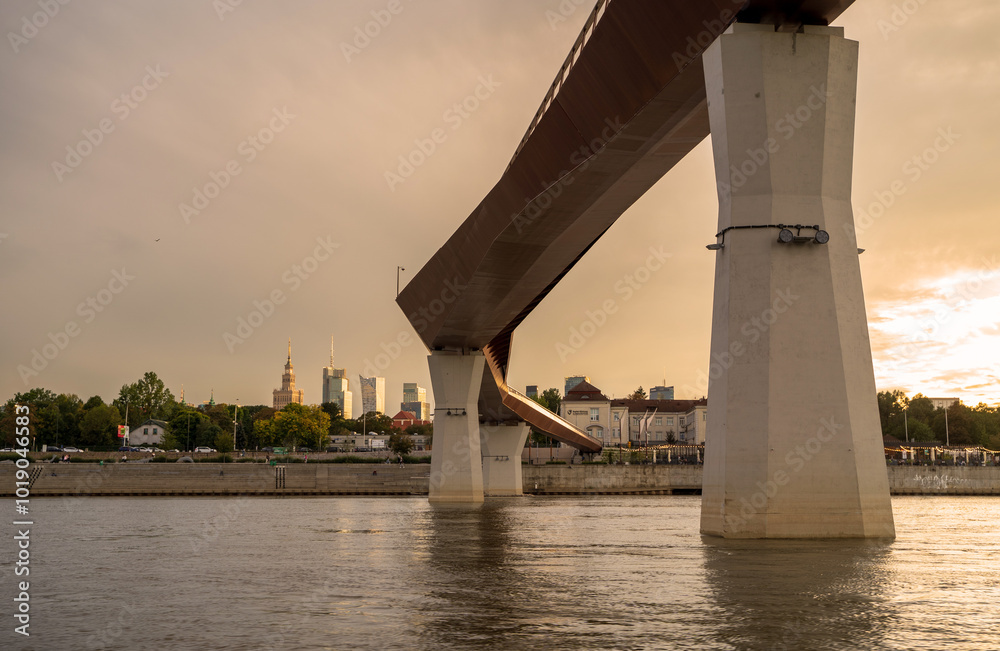 A modern pedestrian bridge with white pillars and a reddish-brown deck stretches across a river in Warsaw, Poland. The setting sun casts a warm glow on the city skyline