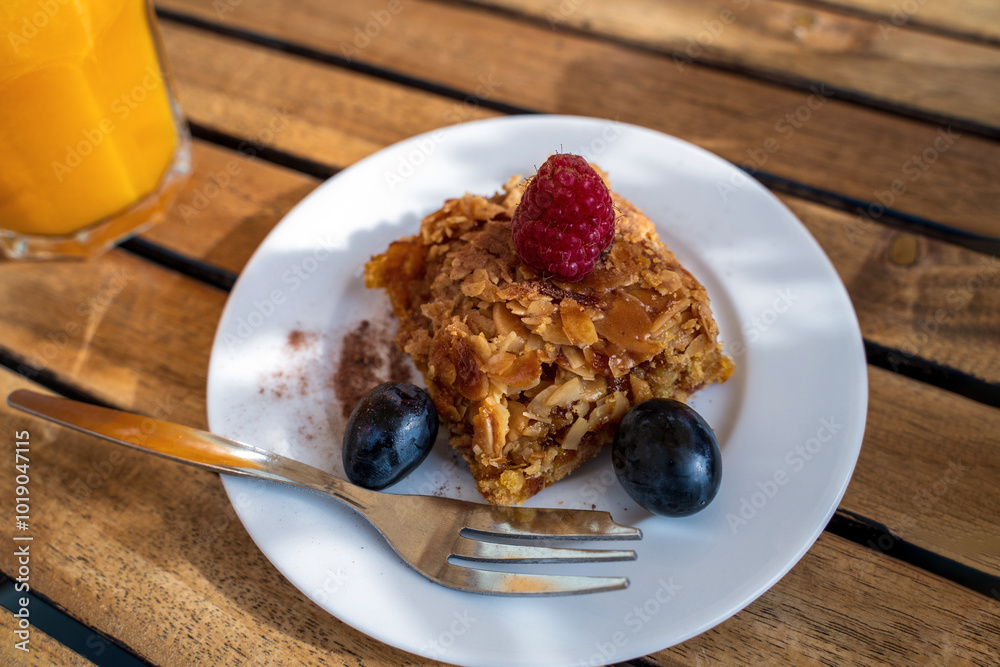 A delicious-looking slice of orange cake sits on a white plate, adorned with a raspberry and surrounded by blueberries. A glass of orange juice complements the sweet treat on a wooden table.