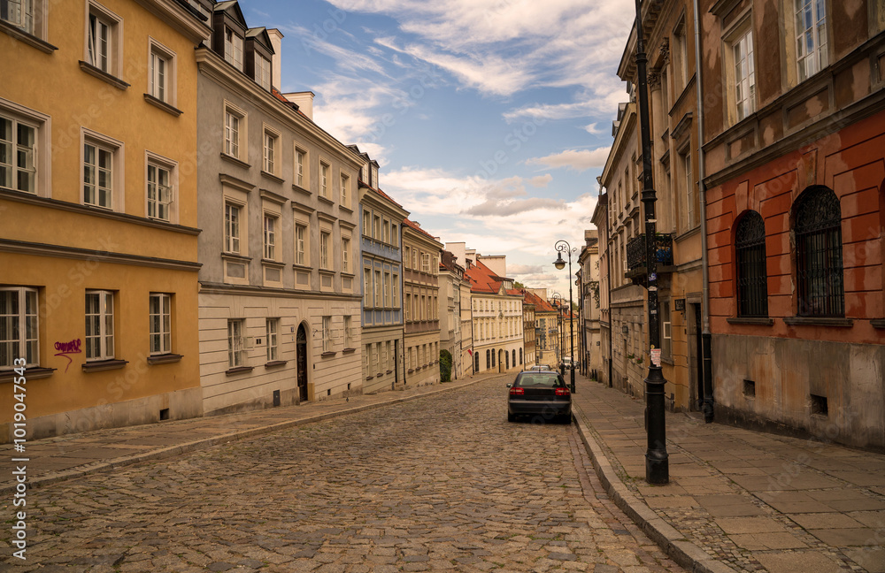 A narrow, cobblestone street slopes gently uphill in Warsaw's Old Town. Colorful historic buildings line both sides, creating a charming and quaint atmosphere.