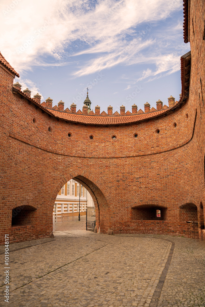 A beautiful view of a historic brick archway leading to a cobblestone path, under a clear blue sky with wispy clouds in Warsaw old town