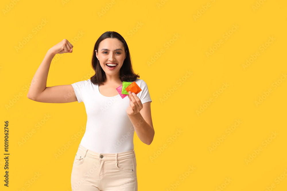Beautiful young woman with condoms showing muscles on yellow background. Safe sex concept Beautiful young woman with condoms showing muscles on yellow background. Safe sex concept
