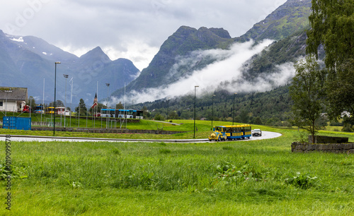 Olden, Norway 15 Aug, 2024 - A tourists's train chugs along a winding road, surrounded by lush green fields and towering mountains shrouded in mist