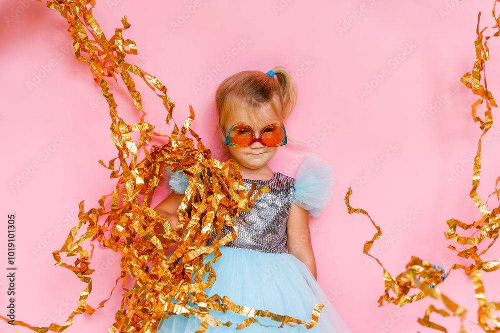 Girl with long hair in festive paper hat and dressy dress on pink background tossing gold confetti Girl with long hair in festive paper hat and dressy dress on pink background tossing gold confetti