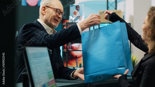 Obraz na plátně Checkout staff selling clothing items to senior customer in shopping mall, preparing clothes in a paper bag for client