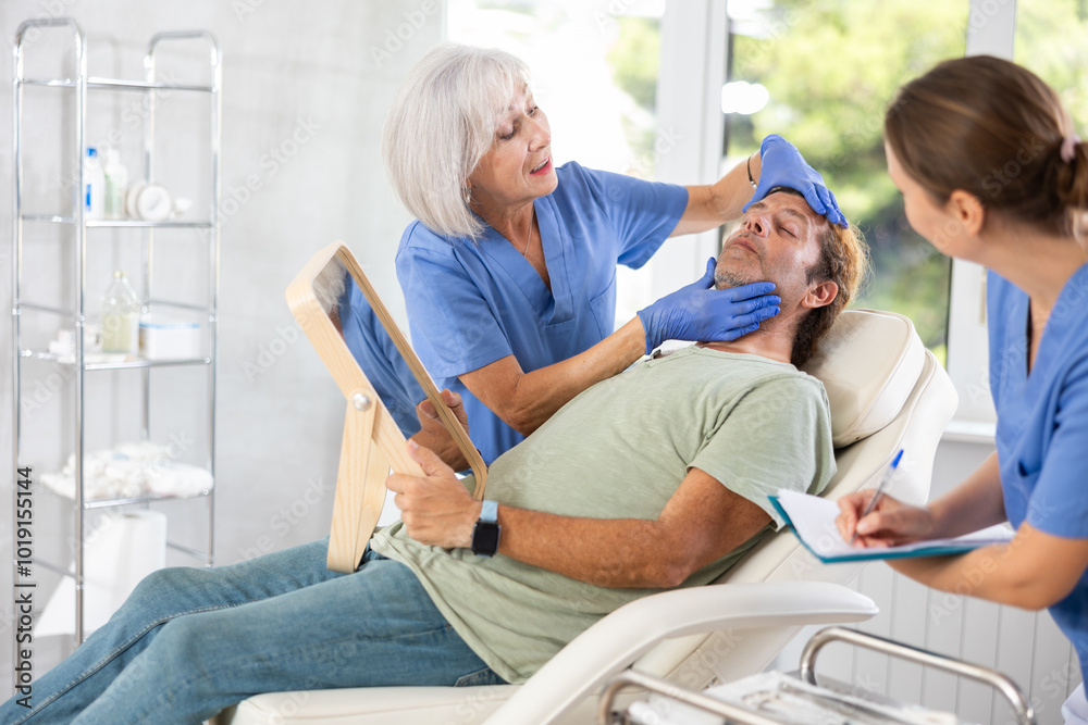 Obraz premium Beautician with nurse talking with patient before injection treatment, while client observing herself in mirror.
