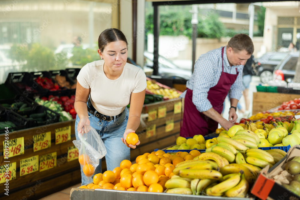Obraz premium Young female shopper selects and buys fresh oranges in a supermarket