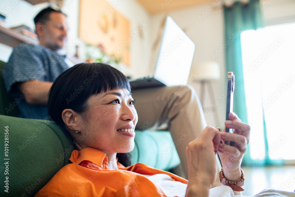 Happy Asian woman using smartphone while relaxing at home with man working on laptop in background