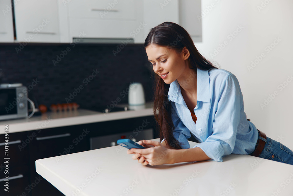 Woman in blue shirt sitting on kitchen counter looking at phone in domestic setting Woman in blue shirt sitting on kitchen counter looking at phone in domestic setting