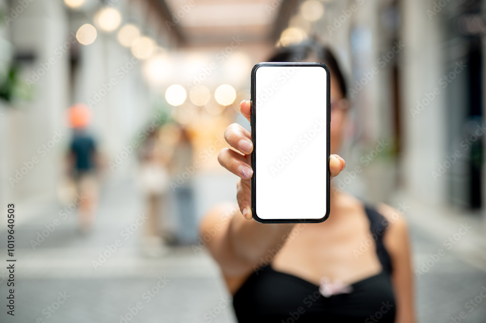 A woman showing a smartphone to the camera while standing in a market or mall. A woman showing a smartphone to the camera while standing in a market or mall.