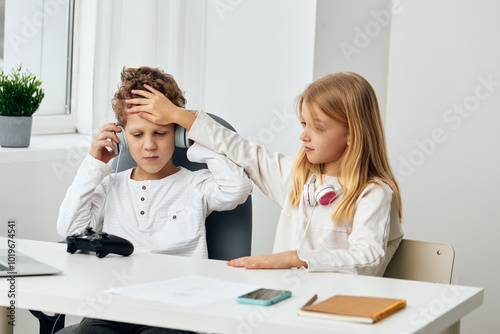 Happy Caucasian boy and girl studying elearning at home, sitting together at the living room table They are using their laptops, wearing headphones, and typing on their phones while playing