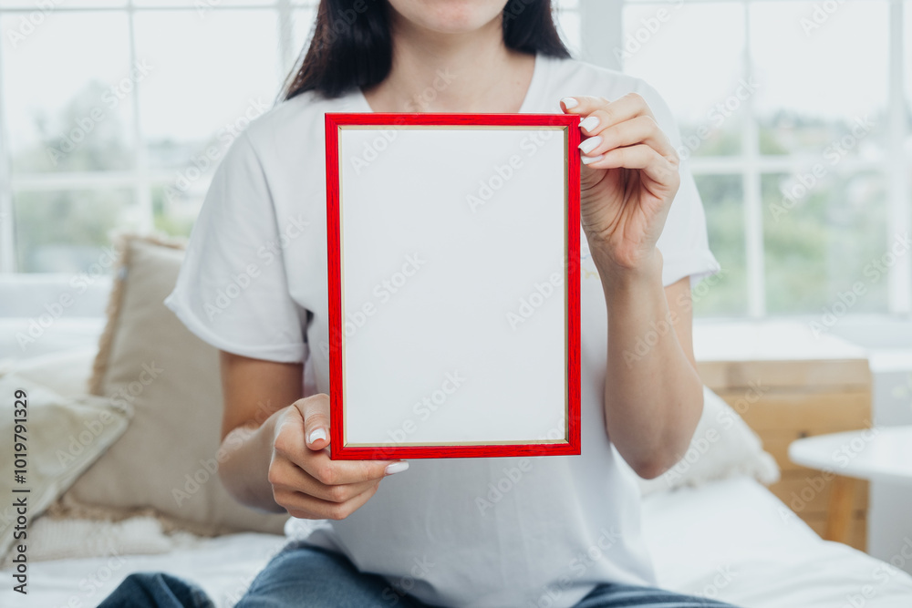 A person holding a blank red frame while sitting on a cozy bed in a bright room A person holding a blank red frame while sitting on a cozy bed in a bright room