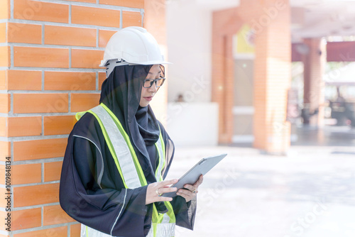 A beautiful Asian Muslim construction engineer in safety vest and Abaya niqab traditional clothes standing and use tablet computer on brick block building wall background.