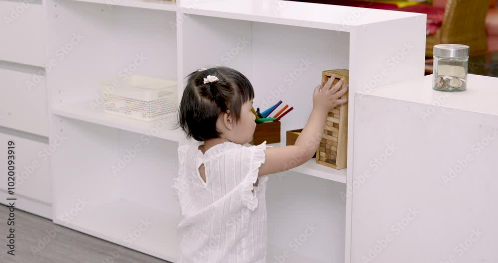 Asian toddler reaching for wooden blocks on shelf, involved in independent play. Scene highlights child’s curiosity and focus on hands-on learning through exploration in home environment. Asian toddler reaching for wooden blocks on shelf, involved in independent play. Scene highlights child’s curiosity and focus on hands-on learning through exploration in home environment.