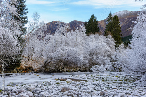 A frozen river meanders through a forest covered in snow, with large rocks lining the riverbed and mountains in the background.