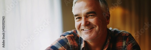 A man with a smile on his face is sitting in front of a window. He is wearing a plaid shirt and he is happy
