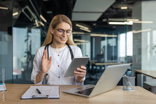 Smiling female doctor in office conducts virtual consultation using tablet, laptop. Medical professional embracing technology for effective communication, remote healthcare solutions.