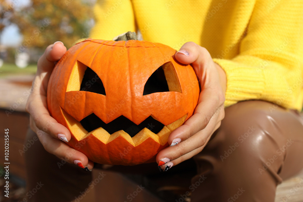 Young woman in yellow sweater holding orange carved Halloween pumpkin in her hands, no face, unrecognizable. Halloween traditional smiling jack-o-lantern. Person sitting in autumn park outdoor.