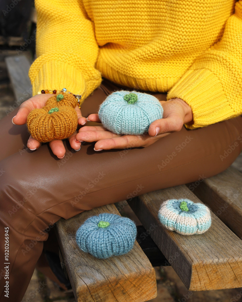 Woman's hands holding three knitting pumpkins orange and blue, two more blue and white are on park bench. Person wearing yellow sweater and brown leather pants. Fall autumn concept.