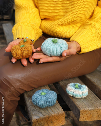 Woman's hands holding three knitting pumpkins orange and blue, two more blue and white are on park bench. Person wearing yellow sweater and brown leather pants. Fall autumn concept.