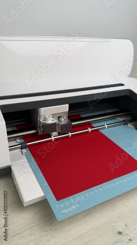 Vertical video of plotter in process of cutting details on red cardboard on blue mat. DIY manual paper craft, making gift for family. Creative project. Cutting machine works, digital flatbed cut out 