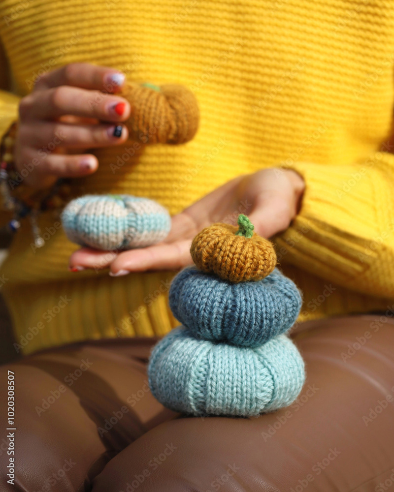 Three colorful handmade knitting pumpkins in front and two others in woman's hands on background. Fall autumn concept. Person unrecognizable, wearing yellow sweater, brown leather pants.
